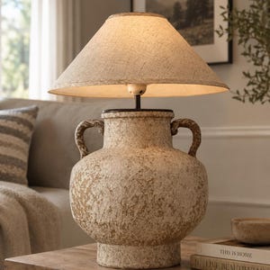 Puede incluir: Una lámpara de mesa con una base de cerámica texturizada en tonos beige y marrón, con forma de ánfora y dos asas laterales. La lámpara cuenta con una pantalla cónica de tela en color claro que emite una luz cálida desde el interior.