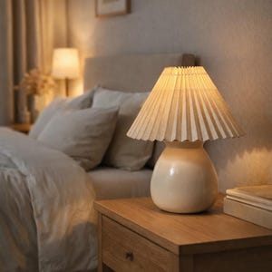 Może przedstawiać: Kremowa lampka nocna z plisowanym abażurem stoi na drewnianym stoliku nocnym. Lampa ma zaokrągloną podstawę i emituje ciepłą poświatę. W sypialni znajduje się łóżko z poduszkami i obraz w ramie.