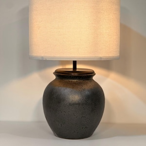 Könnte beinhalten: Dekorative Tischlampe mit einem strukturierten, dunkelgrauen Keramikfuß und einem beigefarbenen Stoffschirm. Die Lampe hat einen runden, urnenförmigen Sockel mit braunem Rand und einen zylindrischen Lampenschirm.