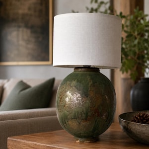 Op de afbeelding: Een tafellamp met een bolvormige, getextureerde keramische voet met een gevlekte groene en bruine glazuurlaag. De lamp is voorzien van een cilindrische, gebroken witte stoffen kap die een neutraal contrast vormt met de organische aardetinten.