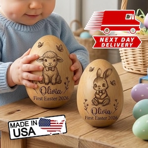 Peut inclure: Deux œufs de Pâques en bois, l'un avec un agneau et l'autre avec un lapin, gravés de "Olivia First Easter 2026". Les œufs sont sur une surface en bois, avec un autocollant "Made in USA" visible.