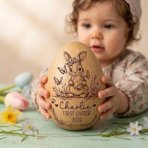 Peut inclure: Un œuf de Pâques en bois avec un motif gravé d'un lapin tenant un œuf, entouré de papillons et de fleurs. L'œuf porte l'inscription "Charlie" et "FIRST EASTER 2026".