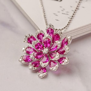 May include: A silver necklace with a floral pendant. The pendant is made of pink gemstones and small white diamonds. The necklace is presented on a white surface, with a monochrome image visible in the background.