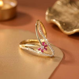 May include: A gold-toned open ring featuring a V-shaped design adorned with clear pavé-set crystals along the bands. A small flower-shaped accent sits at the centre, crafted from six baguette-cut deep pink rubies surrounding a single clear crystal.