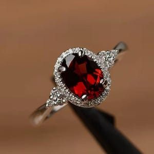 May include: A silver-toned ring featuring a large, oval-cut deep red garnet gemstone at the center. The garnet is surrounded by a halo of small, clear, round-cut stones, with additional small clear stones set along the band on both sides.