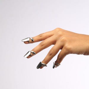 May include: A set of four silver-toned metal nail rings designed to fit over the fingertips. Each ring features a smooth, elongated, curved plate that extends over the nail bed, secured by a thin, adjustable band that wraps around the finger.