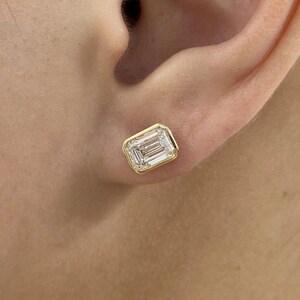 Könnte beinhalten: Einzelner Ohrstecker mit Smaragdschliff-Diamanten und Goldfassung. Der klare Diamant ist rechteckig mit sichtbaren Facetten. Der Ohrring befindet sich vor dem Hintergrund eines Ohres.
