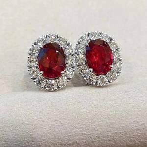 May include: A pair of oval-cut ruby earrings. Each earring features a vibrant red ruby surrounded by a halo of small, sparkling white diamonds. The earrings are set in a silver or white gold setting, creating a classic and elegant look.