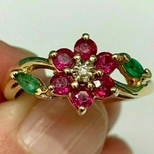Puede incluir: Un anillo de oro con un diseño floral central compuesto por seis rubíes rojos redondos que rodean un pequeño diamante transparente. La banda incluye dos esmeraldas verdes de talla marquesa a cada lado del centro floral, en oro pulido.
