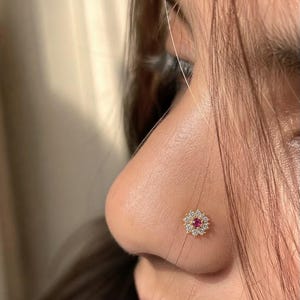 May include: A gold nose stud featuring a central round red gemstone surrounded by a halo of small clear white stones arranged in a flower-like pattern. The jewelry is positioned on the side of a nose, showcasing the circular floral design.