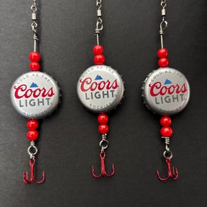 May include: Three handmade fishing lures. Each lure features a silver Coors Light bottle cap with red text and a red treble hook. Red beads are strung between the bottle cap and the hook. The lures are suspended from a thin wire.