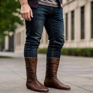 Può includere: Stivali in pelle marrone con un design al ginocchio. Gli stivali presentano un dettaglio stringato e sono indossati con jeans blu. Gli stivali sono di colore marrone medio e hanno una suola piatta.