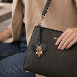 May include: A black textured leather handbag featuring a gold-toned zipper and a heart-shaped black leather bag charm. The charm displays a detailed illustration of a golden retriever wearing a royal crown and a red velvet cape with white fur trim.