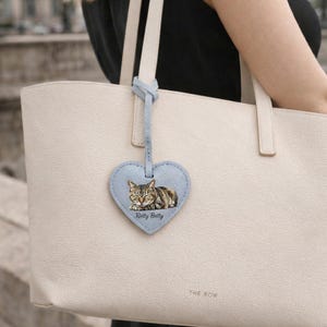 May include: A cream-colored tote bag with a light blue heart-shaped leather tag attached to the handle. The tag features a printed image of a sleeping tabby cat and the text Retty Betty printed in black ink below the cat illustration.