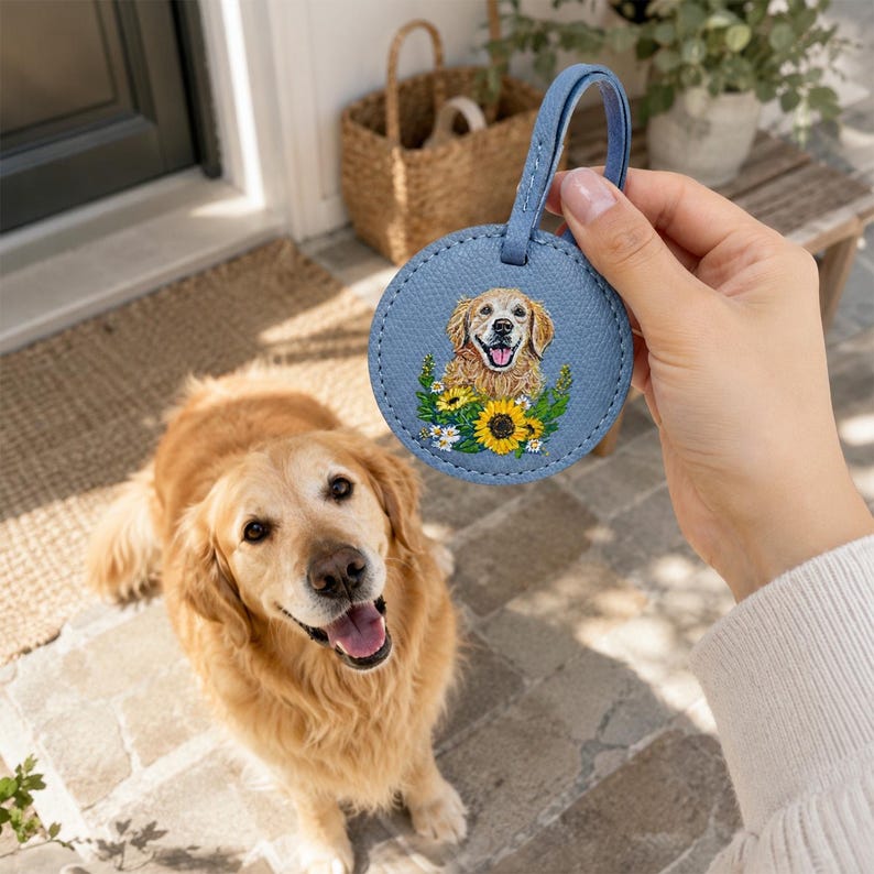 May include: A round, light blue leather luggage tag featuring a detailed illustration of a golden retriever dog surrounded by yellow sunflowers and small white daisies. The tag has a matching blue leather strap attached to the top circular edge.