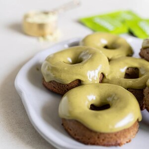 Pu&ograve; includere: Un piatto di ceramica bianca contiene diverse ciambelle cotte al forno con una base marrone chiaro e una glassa verde chiaro e liscia. Un misurino di polvere bianca e una bustina verde sono sullo sfondo.