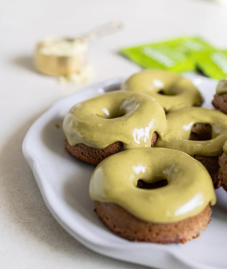 Pu&ograve; includere: Un piatto di ceramica bianca contiene diverse ciambelle cotte al forno con una base marrone chiaro e una glassa verde chiaro e liscia. Un misurino di polvere bianca e una bustina verde sono sullo sfondo.