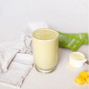 Pu&ograve; includere: Una tazza di vetro trasparente piena di un frullato giallo chiaro. Un misurino bianco con polvere gialla, bustine verdi e pezzi di mango tagliati sono accanto alla tazza. Un tovagliolo bianco &egrave; a sinistra.