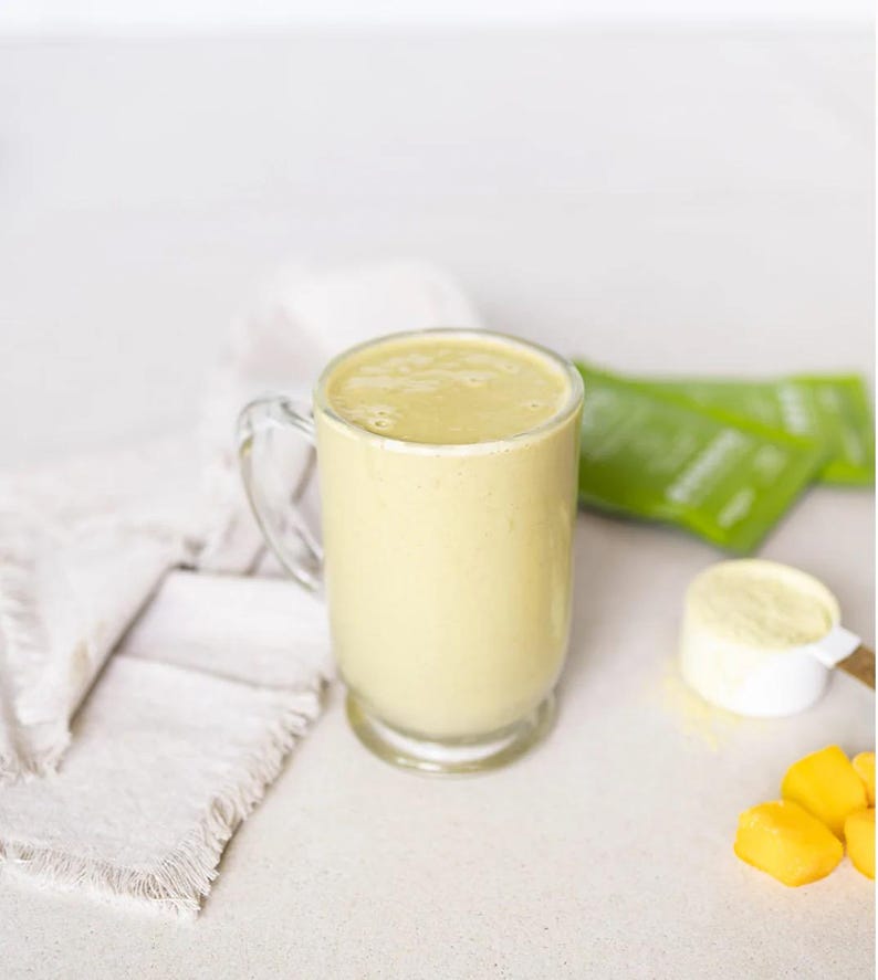 Pu&ograve; includere: Una tazza di vetro trasparente piena di un frullato giallo chiaro. Un misurino bianco con polvere gialla, bustine verdi e pezzi di mango tagliati sono accanto alla tazza. Un tovagliolo bianco &egrave; a sinistra.