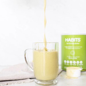 Pu&ograve; includere: Un frullato giallo viene versato in una tazza di vetro trasparente. Un contenitore verde e bianco con la scritta "HABITS" &egrave; sullo sfondo, insieme a un misurino di polvere. Un panno beige &egrave; sulla superficie.