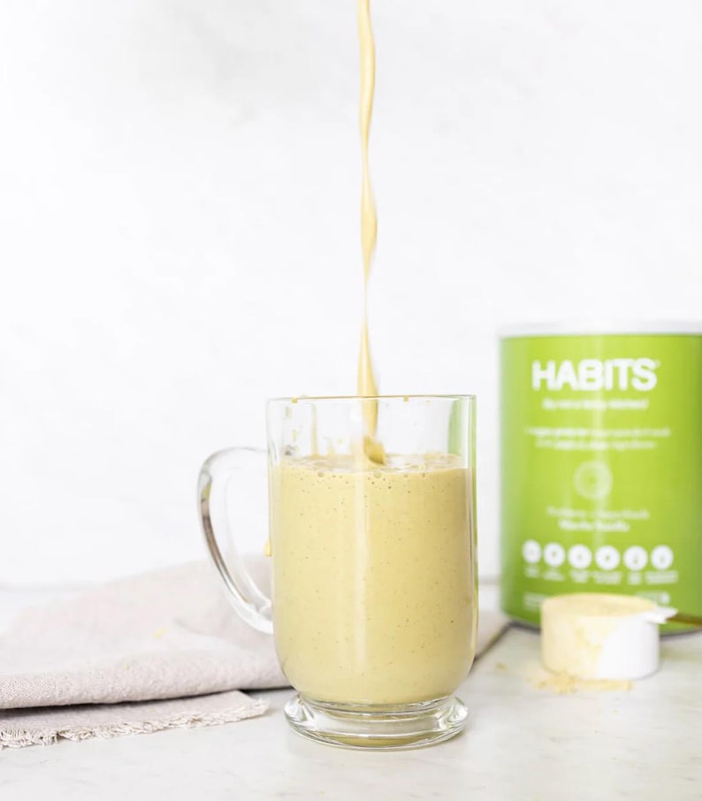 Pu&ograve; includere: Un frullato giallo viene versato in una tazza di vetro trasparente. Un contenitore verde e bianco con la scritta "HABITS" &egrave; sullo sfondo, insieme a un misurino di polvere. Un panno beige &egrave; sulla superficie.