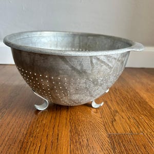May include: A round, silver-coloured metal colander featuring a wide rim and a series of small, evenly spaced drainage holes across its bowl. The kitchen tool is supported by three curved metal feet attached to the base, providing stability.
