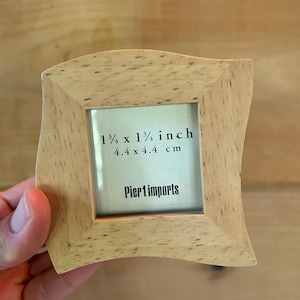 May include: A square light-colored wooden picture frame with a wavy edge design. The frame contains a display card with the text 1 3/4 x 1 3/4 inch, 4.4 x 4.4 cm, and Pier 1 imports printed in black ink on a light background.