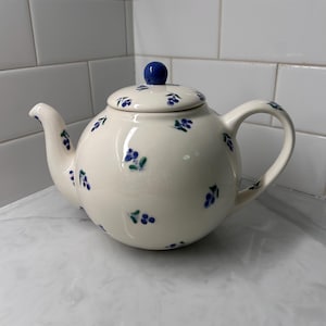 May include: Cream-colored ceramic teapot with a rounded body, spout, and handle. The teapot features a lid with a blue knob and is decorated with small blue floral patterns. The teapot is sitting on a marble countertop.