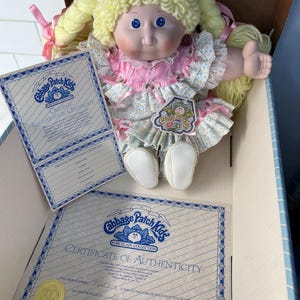 May include: A Cabbage Patch Kids porcelain doll with blonde yarn hair styled in pigtails, wearing a pink and floral dress. Includes a registration certificate and certificate of authenticity.
