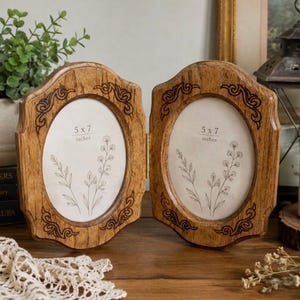 May include: A hinged double photo frame made of light brown wood with a visible grain. Each oval-shaped frame features dark decorative scrollwork at the top and bottom corners. The inserts clearly display the text 5 x 7 inches with floral line art.