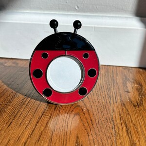 Op de afbeelding: Een ronde metalen fotolijst in de vorm van een lieveheersbeestje met een glanzend rood lichaam, zwarte stippen en een zwart kopgedeelte. De lijst heeft twee zwarte voelsprieten aan de bovenkant en een centrale ronde opening voor een foto.