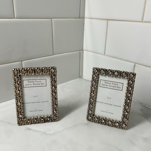 May include: Two rectangular picture frames with metallic bronze-toned borders embedded with clear crystals. Each frame contains a white insert labeled Three Piece Jewel Frame Set, 2x3, genuine austrian crystals, by Sheffield Home.