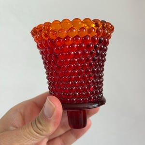 May include: A glass hobnail votive candle holder featuring a vibrant gradient color scheme that transitions from a deep, saturated red at the base to a bright, warm orange at the flared rim, with a distinct textured surface of raised glass beads.