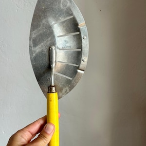 May include: A vintage Foley kitchen tool featuring a semi-circular metal blade with five parallel slots and a bright yellow cylindrical wooden handle. The silver-toned metal surface shows signs of wear and includes the embossed brand name Foley.