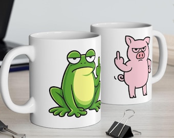 May include: Two white ceramic mugs with humorous cartoon illustrations. One mug features a green frog with a yellow belly, and the other depicts a pink pig. Both characters are giving the middle finger. The mugs have a standard handle.