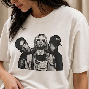 May include: A cream-colored short-sleeve t-shirt featuring a black and white graphic print of the band Nirvana. The print shows three band members, with the central figure wearing white oval sunglasses and a patterned shirt with the text XES.