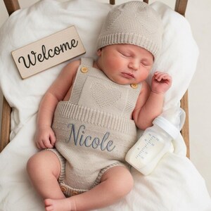 Puede incluir: Un recién nacido durmiendo, vestido con un pelele de punto beige con el nombre "Nicole" bordado en azul. El bebé también lleva un gorro a juego. En la imagen también hay un biberón y un cartel de madera con la palabra "Welcome".