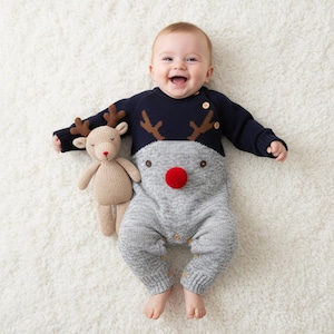 May include: A baby wearing a navy blue and gray reindeer-themed knit romper with a red pom-pom nose and brown antlers. The baby is smiling and lying on a white fluffy surface next to a stuffed reindeer toy.