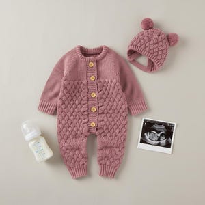 May include: A dusty rose knitted baby outfit set, including a romper with wooden buttons and a matching hat with pom-poms. A baby bottle and an ultrasound image are also in the image.