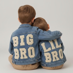 May include: Two children wearing matching denim jackets with the words "BIG BRO" and "LIL BRO" appliqued on the back. The jackets are light blue with beige lettering. The children are embracing each other.