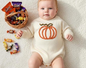 May include: A cream-coloured long-sleeve baby romper featuring a central orange pumpkin embroidery with a green stem. Beside the baby is a small woven wicker basket filled with various colourful candy wrappers and several loose sweets.