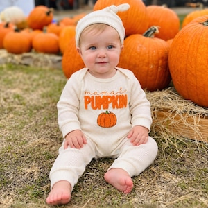 Peut inclure: Une grenouillère à manches longues en tricot gaufré couleur crème avec un bonnet assorti. La grenouillère présente le texte brodé orange 'mama's PUMPKIN' au-dessus d'un petit motif de citrouille orange. Le tissu a une texture gaufrée.