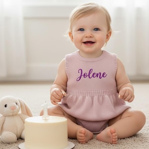 Pode incluir: Um bebê sorrindo usando um macacão de malha rosa com o nome "Jolene" em escrita roxa. Um pequeno bolo branco com uma vela número um está na frente do bebê, ao lado de um coelhinho de pelúcia.