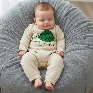 Puede incluir: Un bebé dormido con un pelele de punto beige con un diseño de tortuga verde en la parte delantera. El bebé descansa sobre un gran puf gris de felpa. El pelele tiene mangas largas y cierres de botón a presión en las piernas.