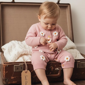May include: A baby in a pink knit romper adorned with white daisy appliqués sits in an open vintage suitcase. The romper has wooden buttons and is paired with a matching cardigan. A tag reads "Sweet One".
