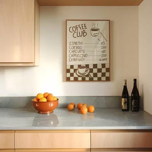 May include: A framed wall print titled COFFEE CLUB with a list of coffee drinks and a checkered pattern at the bottom. The text reads: ESPRESSO YES, CORTADO ICED, CHAI LATTE HOT, CAPPUCCINO ICED, EXTRA SHOT ALWAYS. A hand-drawn coffee cup is included.