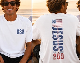 May include: A white short-sleeve t-shirt featuring the text USA in blue on the front left chest. The back displays a vertical graphic with an American flag, the word JESUS, the text GOD SHED HIS GRACE ON THEE, and 1776 2026 250 in red and blue.