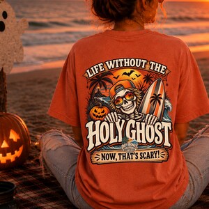 May include: A burnt orange t-shirt featuring a graphic on the back of a skeleton wearing sunglasses and a baseball cap, holding a surfboard. The design includes a pumpkin, palm trees, a sunset, and the text: LIFE WITHOUT THE HOLY GHOST NOW, THAT'S SCARY!