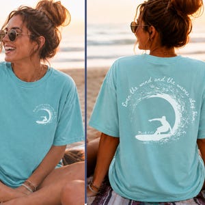 May include: A light blue t-shirt with a white graphic of a surfer riding a wave. The text "Even the wind and the waves obey Him" is printed on the back. The shirt is a comfortable, casual style.