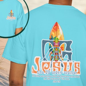 May include: A light blue t-shirt with a colorful graphic featuring a surfboard, tiki masks, and the word "Jesus." The text "the OG wave walker" and a Bible verse are also printed on the shirt.